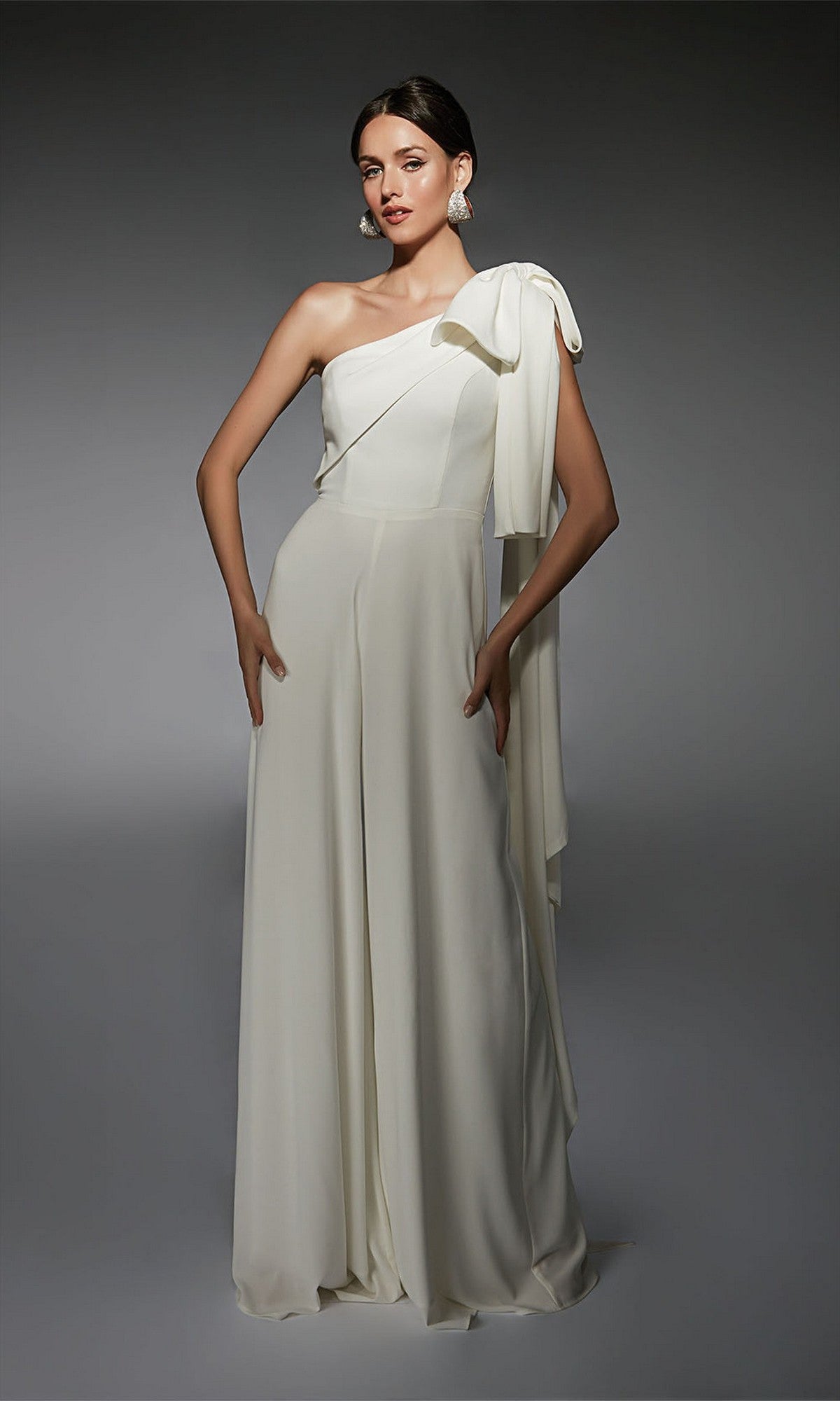 A woman models the Alyce 70065 long formal jumpsuit—a white, floor-length, one-shoulder outfit with a large bow detail—offering a chic cocktail dress alternative against a plain gray background.